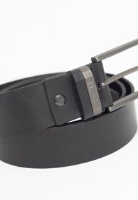 Cinturón de cuero negro con una textura suave, presenta una hebilla metálica con dos púas y un logotipo grabado. Diseño sencillo y clásico.