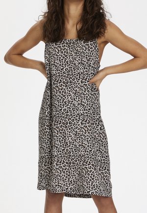 Woman wearing a sleeveless, knee-length dress with brown and black leopard print, hands placed on hips, against a white background.