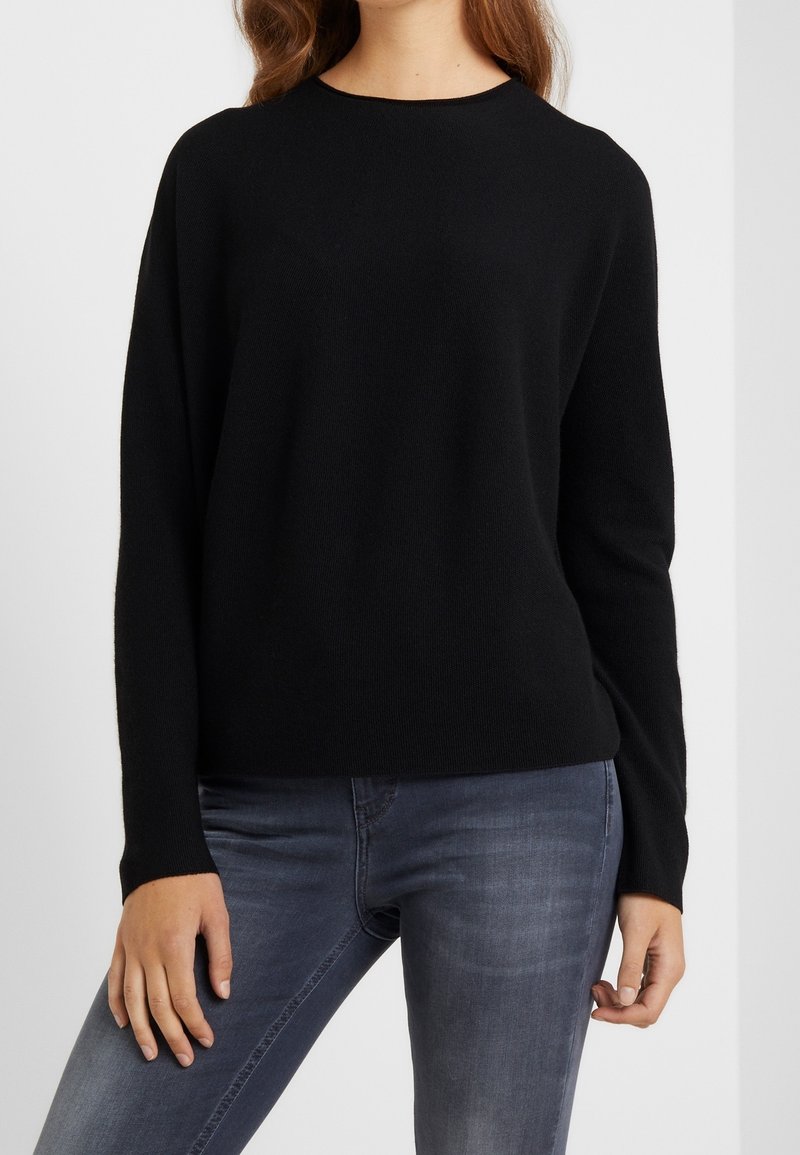 Femme portant un pull noir uni à manches longues associé à un jean bleu délavé, visible du cou aux hanches sur un fond uni.