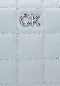 Světle modrá prošívaná kožená taška s stříbrným logem "CK". Má výrazné švy a hladkou texturu, která zdůrazňuje moderní design.