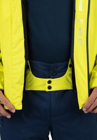 Chaqueta impermeable amarilla con acentos negros, combinada con pantalones azul marino. Presenta botones y un forro interior texturizado.