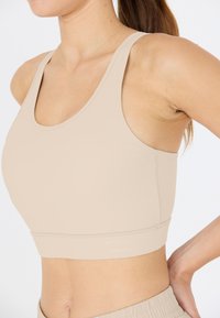 Urheilu-rintaliivit beige värissä, valmistettu sileästä ja joustavasta kankaasta. Ominaisuuksiin kuuluvat leveät hihnat, pyöreä pääntie ja huomaamattomat ompeluyksityiskohdat helman reunassa.