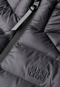 Giacca nera imbottita con sezioni trapuntate orizzontali, dotata di una cerniera visibile e del logo "SUPERDRY" ricamato con filo scuro.