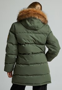 Olivno zeleni napihani plašč z snemljivim rjavim krznom podloženim kapuco, rebrastimi manšeti in teksturiranim prešitim dizajnom. Ima logotip na rokavu.