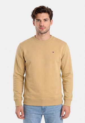 Giovane uomo con capelli corti castani che indossa una felpa beige e jeans azzurri chiari, in piedi davanti a uno sfondo bianco semplice.