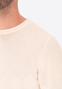 Primo piano del collo e della spalla di un uomo che indossa un maglione lavorato a maglia di un leggero beige con un collo rotondo a coste.
