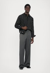 Man in een zwart geblokt lang shirt met lange mouwen, grijze wijde broek, zwarte riem, zwarte schoenen en houdt een kleine zwarte schoudertas vast.