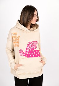 Beige hoodie with a large pink tiger graphic, stars, and orange text. Features a front pocket and an oversized hood. Cotton material.