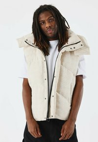 Beige gewatteerde vest met een ruitpatroon, voorzien van een hoge kraag, rits en drukknopen. Gedragen over een wit T-shirt tegen een effen achtergrond.