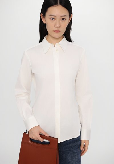 White, collared button-up shirt with long sleeves. Smooth fabric, fitted design. Brown handbag held in hand, with a cut-out handle.