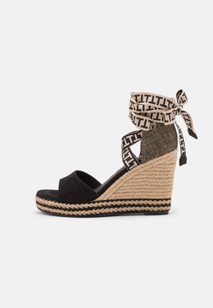 Sorte ruskind wedge sandal med en flettet jute hæl, der har en mønstret stof ankelrem bundet på siden. Sålen har sorte detaljer.