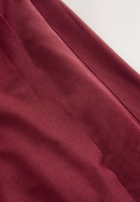 Nærbillede af glat mahognifarvet stof med en synlig søm og lette folder på en lys baggrund.