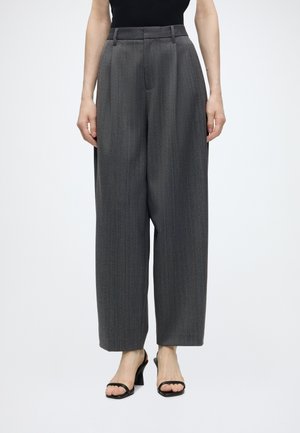 Femme portant un pantalon gris à fines rayures, taille haute et jambes larges, un top noir sans manches et des talons ouverts noirs, debout sur fond blanc.