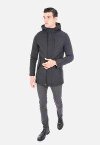 Giacca impermeabile nera con cappuccio, chiusura con zip e due tasche frontali, abbinata a pantaloni grigi slim-fit e scarpe nere.