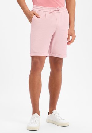 Persoon draagt lichtroze shorts met trekkoord en witte sneakers, staand met één hand in de zak tegen een witte achtergrond.