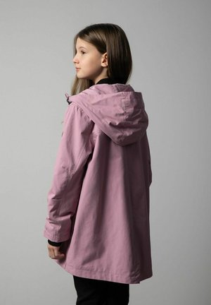 Młoda dziewczyna z długimi brązowymi włosami zwrócona na bok, ubrana w jasnofioletową kurtkę z kapturem i czarne spodnie na jednolitym szarym tle.