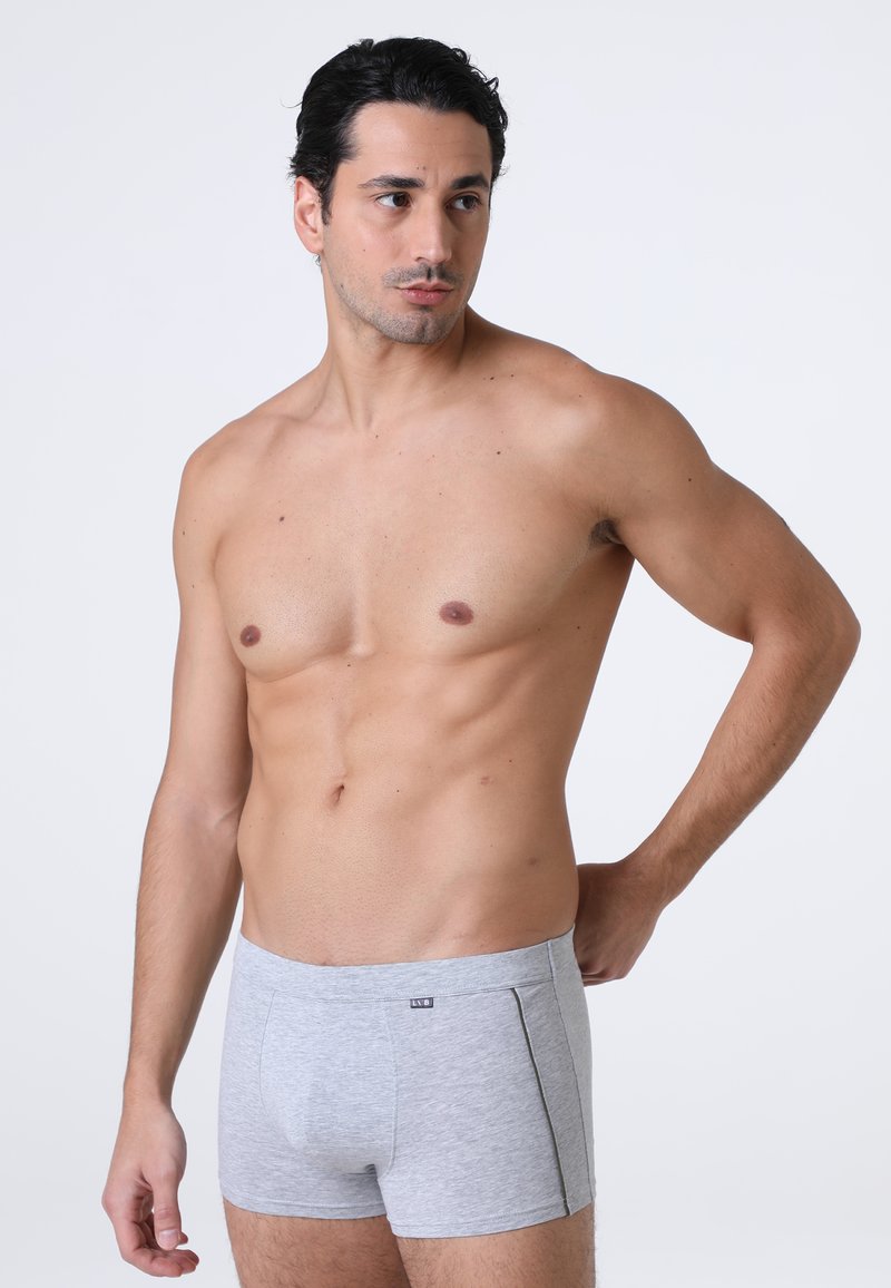 Uomo senza maglietta con capelli scuri che indossa slip grigi chiari, in piedi con una mano dietro la schiena, che guarda di lato su uno sfondo semplice.