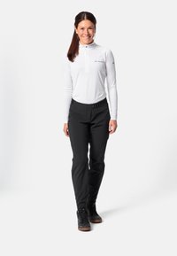 Glunderende vrouw die staat in een witte lange mouwen zip-up top, zwarte broek met riem en zwarte sneakers tegen een effen witte achtergrond.