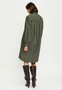 Robe chemise olive, à manches longues, avec un détail froncé au dos, associée à des bottes marron foncé à hauteur de genou. Tissu de texture lisse.