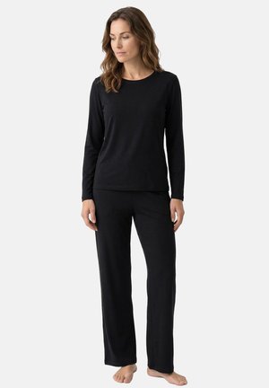 Kobieta stoi boso, ubrana w czarną bluzkę z długim rękawem i czarne szerokie spodnie na jasnym jednolitym tle.