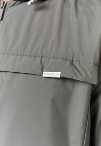 Mørkegrøn letvægtsjakke med lynlås, hætte og en frontlomme. Har et metal logo-plakette med teksten "CLOSURE LDN."