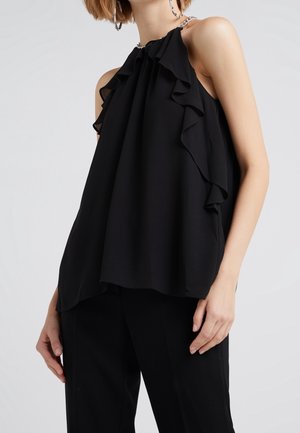 Femme portant un top dos nu noir avec des volants et un détail chaîne, associé à un pantalon noir sur un fond clair uni.