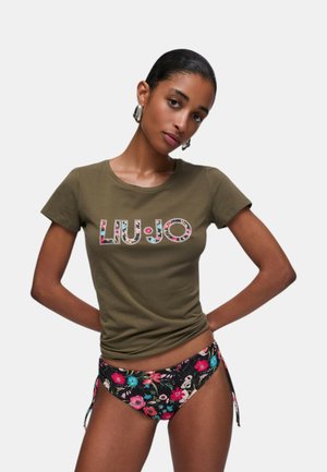 Donna con capelli corti e neri che indossa una maglietta verde oliva con il logo colorato "LIU•JO" e slip bikini neri con fantasia floreale, in piedi con le mani sui fianchi.