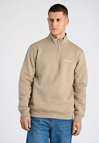 Sudadera beis con cremallera y cuello alto, y puños acanalados. Presenta un pequeño logo en el pecho. Combinada con jeans de denim azul.