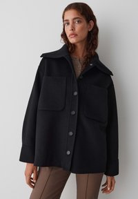 Veste en laine noire avec un col pointu, deux poches avant et des boutons sur le devant. Elle présente une coupe décontractée et une texture douce.