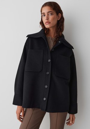 Black wool jacket with a pointed collar, two front pockets, and buttons down the front. Features a relaxed fit and soft texture.