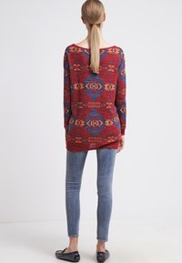 Roter und blauer Pullover mit Muster, weitem Ausschnitt, langen Ärmeln und einem etwas längeren Rückenteil, kombiniert mit eng anliegenden blauen Jeans.