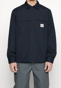 Navy work jacket with a collared neckline, two front pockets, and a logo patch. Made of a smooth fabric with long sleeves and a straight cut.