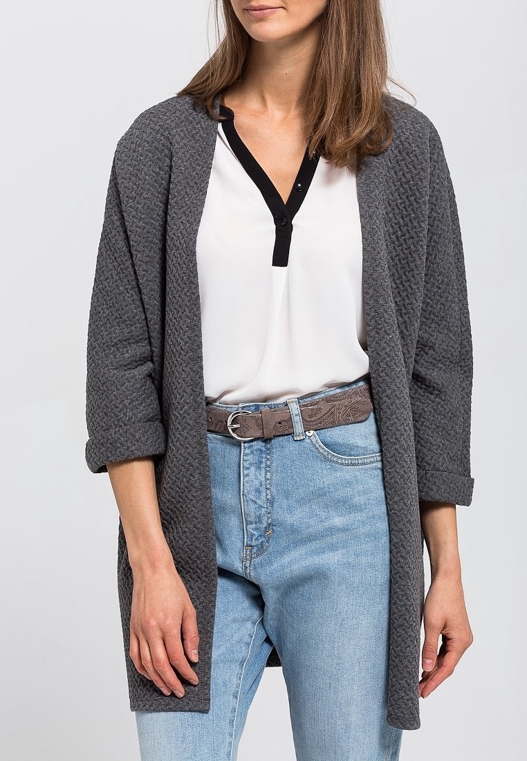 Femme portant un cardigan ouvert texturé gris foncé, une blouse blanche avec un bord noir, un jean bleu clair et une ceinture marron à motifs.