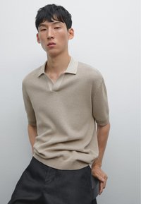 Polo shirt beige lavorato a maglia con colletto corto e motivo testurizzato, abbinato a pantaloni in tessuto scuro, il tutto su uno sfondo neutro.
