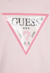 Lys rosa t-skjorte med et stort trekantet paljettmerke med "GUESS" og "U.S.A." i svart, omgitt av rosa perler.