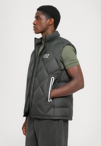 Bărbat îmbrăcat într-o vestă neagră matlasată cu logo-ul EA7, peste un tricou polo de culoare verde măslinie, cu o mână în buzunarul vestei, întorcându-se spre stânga.