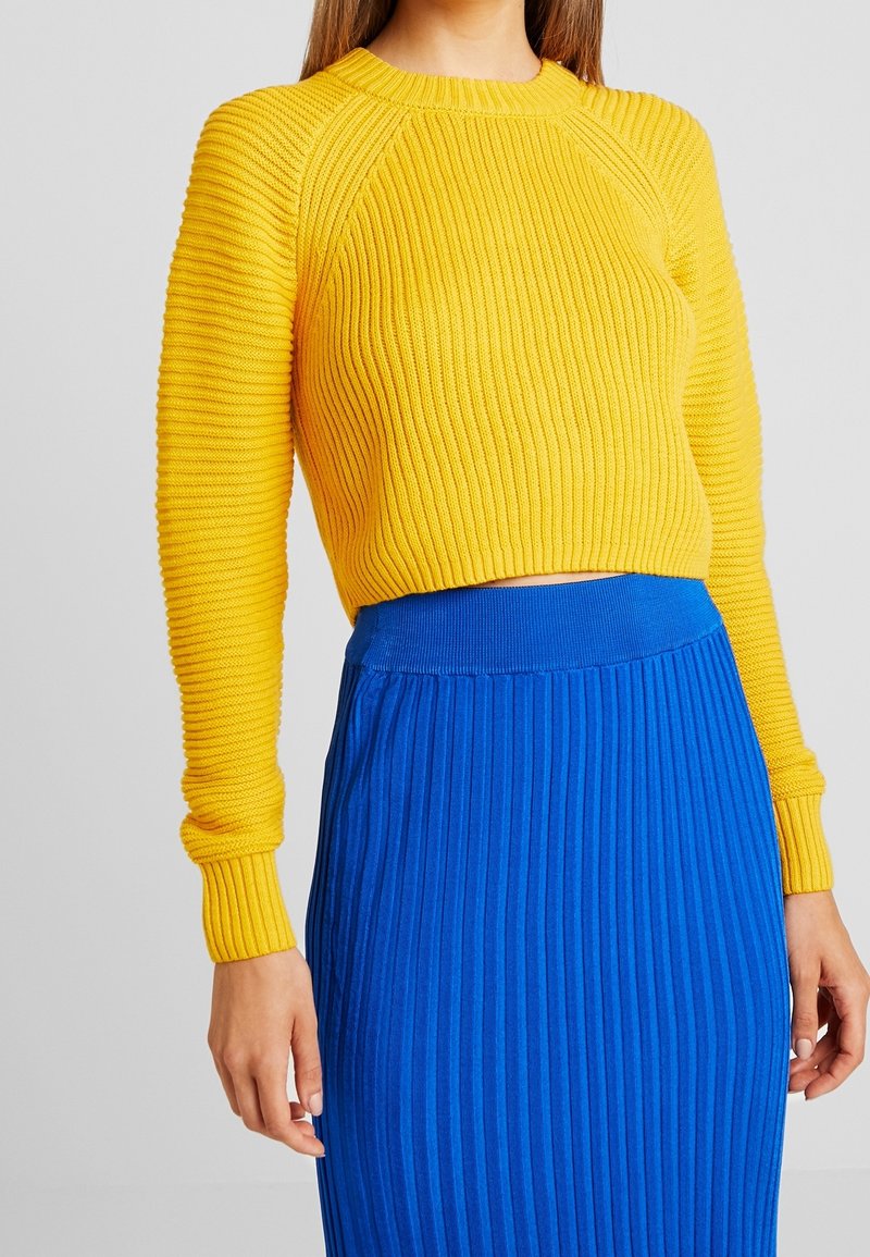 Femme portant un pull jaune côtelé à manches longues et une jupe bleue côtelée taille haute sur un fond uni.