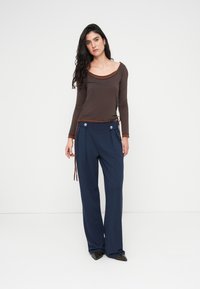 Καφέ μακρυμάνικο τοπ με στρογγυλή λαιμόκοψη και κορδόνια διακόσμησης, συνδυασμένο με μπλε σκούρες φαρδιές παντελόνες με κουμπιά.