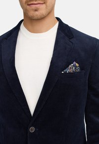 Blazer en velours côtelé marine avec une finition texturée, fermeture à un bouton, et un poche carré à motifs dans des tones de bleu, orange et jaune.