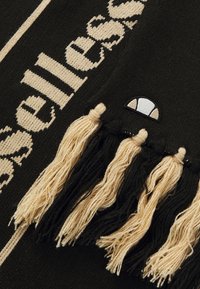 Πλεκτό μαύρο κασκόλ με μπεζ κείμενο και τασιλάκια. Περιλαμβάνει κεντημένο λογότυπο σε ημικυκλικό σχήμα κοντά στην άκρη.