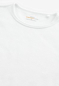 Tricou termic alb cu maneci lungi, textură riglenă, decolteu rotund și etichetă de branding subtilă. Materialul pare moale și elastic.