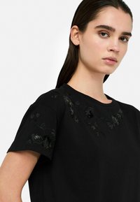 Schwarzes Kurzarm-T-Shirt mit gestickten Blumenmotiven in dunklen Farbtönen entlang des Ausschnitts und der Schultern. Aus glattem Baumwollstoff.
