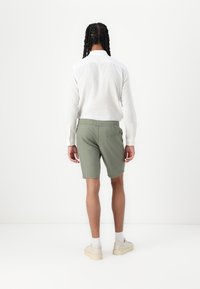 Chemise blanche en lin léger à manches longues associée à un short vert avec une taille élastique et une poche arrière, portée avec des chaussures beiges.