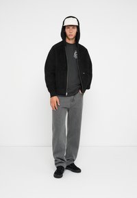 Sort oversized hoodie med en tekstureret overflade, grå T-shirt, løse grå jeans og sorte sneakers. Hvid kasket med flad skygge.