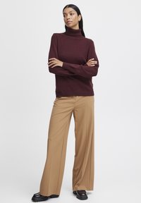 Burgundin رنگی کا ٹرٹleneck sweater جو کہ چوڑے پیروں والی ٹین پینٹس کے ساتھ پہنا گیا ہے۔ یہ لباس ہموار ساخت اور آرام دہ فٹ کا حامل ہے، جس کے ساتھ سیاہ جوتے ہیں۔