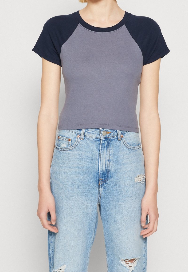 Šedý crop top s námořnickými raglánovými rukávy, vyrobený z měkké látky. Kombinovaný s vysokým pasem ležérními světle modrými džínami s outfitem s roztrhanými akcenty.