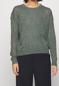 Grön stickad tröja med chevronöppen mönster på ärmar och sidor, bärs med svarta byxor, visad från axlarna till höfterna.