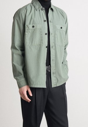 Mann trägt ein hellgrünes Hemd über einem schwarzen Rollkragenpullover, kombiniert mit schwarzen plissierten Hosen und einem Gürtel.