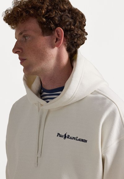 Κρέμα φούτερ με μεγάλη μπροστινή τσέπη, ρυθμιζόμενη κουκούλα και λογότυπο "Polo Ralph Lauren" σε ναυτικό μπλε. Από κάτω, διακρίνεται μια μπλε και άσπρη ριγέ μπλούζα.