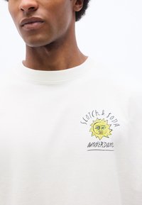 Άνδρας που φοράει λευκό πουκάμισο με μικρό γραφικό ήλιου και κείμενο "Scotch & Soda Amsterdam" στο αριστερό στήθος.